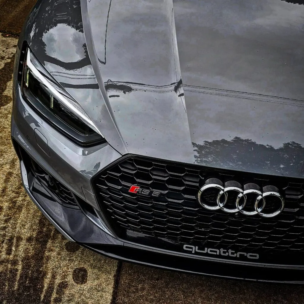 Close-up of a gray Audi RS5 front grille, RS5 badge, and polished hood reflecting trees and sky