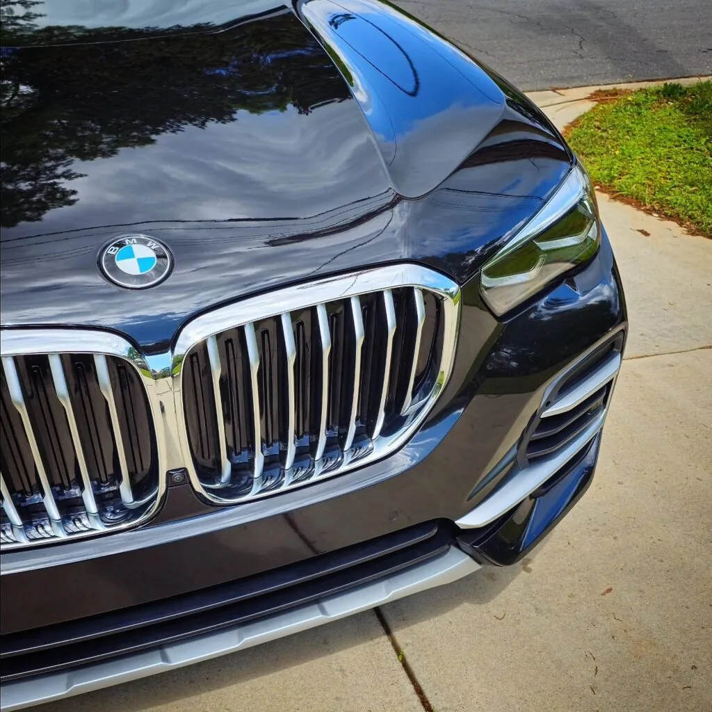 Close-up of a black BMW SUV hood, chrome kidney grille, and headlight with glossy reflective paint in a driveway
