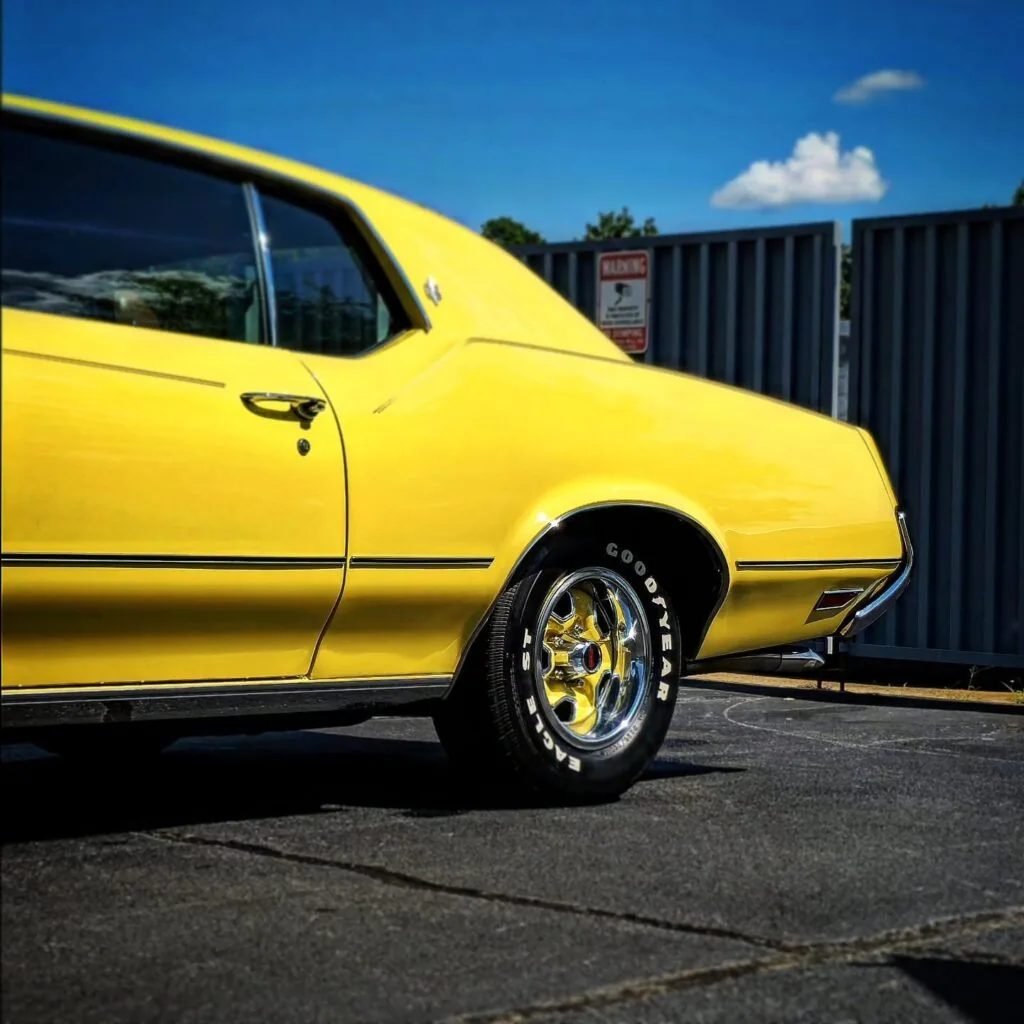 Rear quarter view of a yellow classic Oldsmobile coupe with chrome wheel and white letter Goodyear tire on pavement.