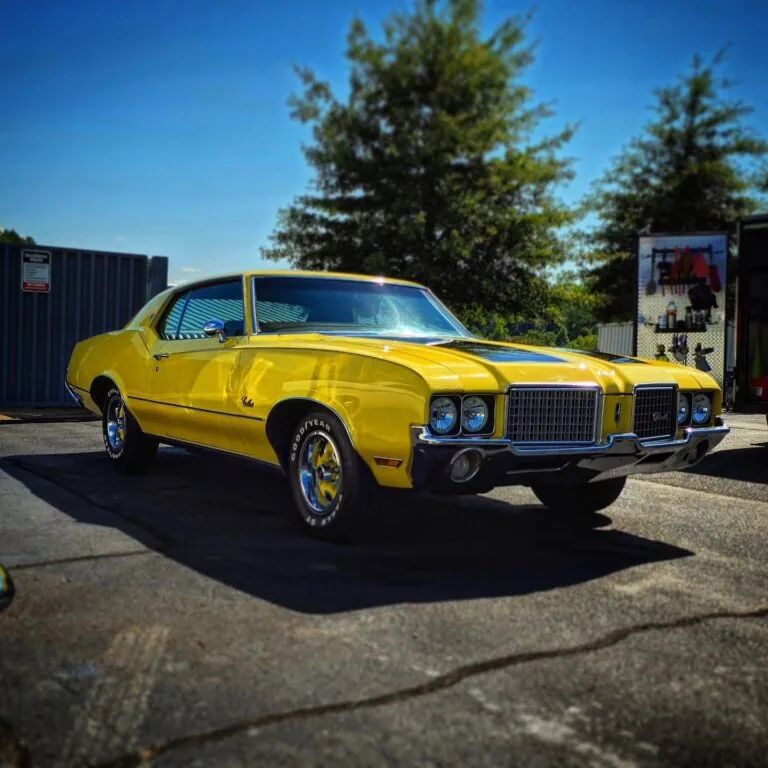 Front quarter view of a yellow Oldsmobile Cutlass with black hood stripes and whitewall lettering tires parked outdoors
