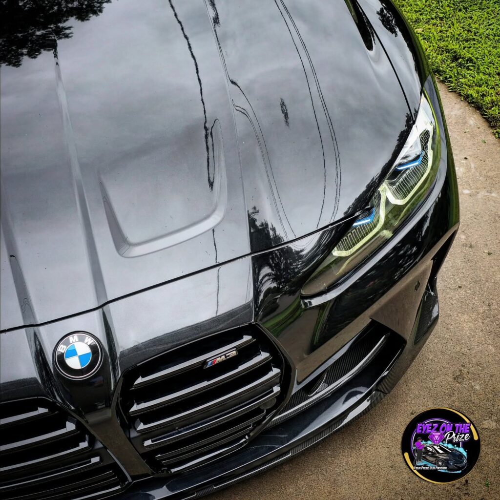 Close-up of a black BMW M3 hood, roundel badge, and kidney grille with a glossy reflective finish on a driveway.