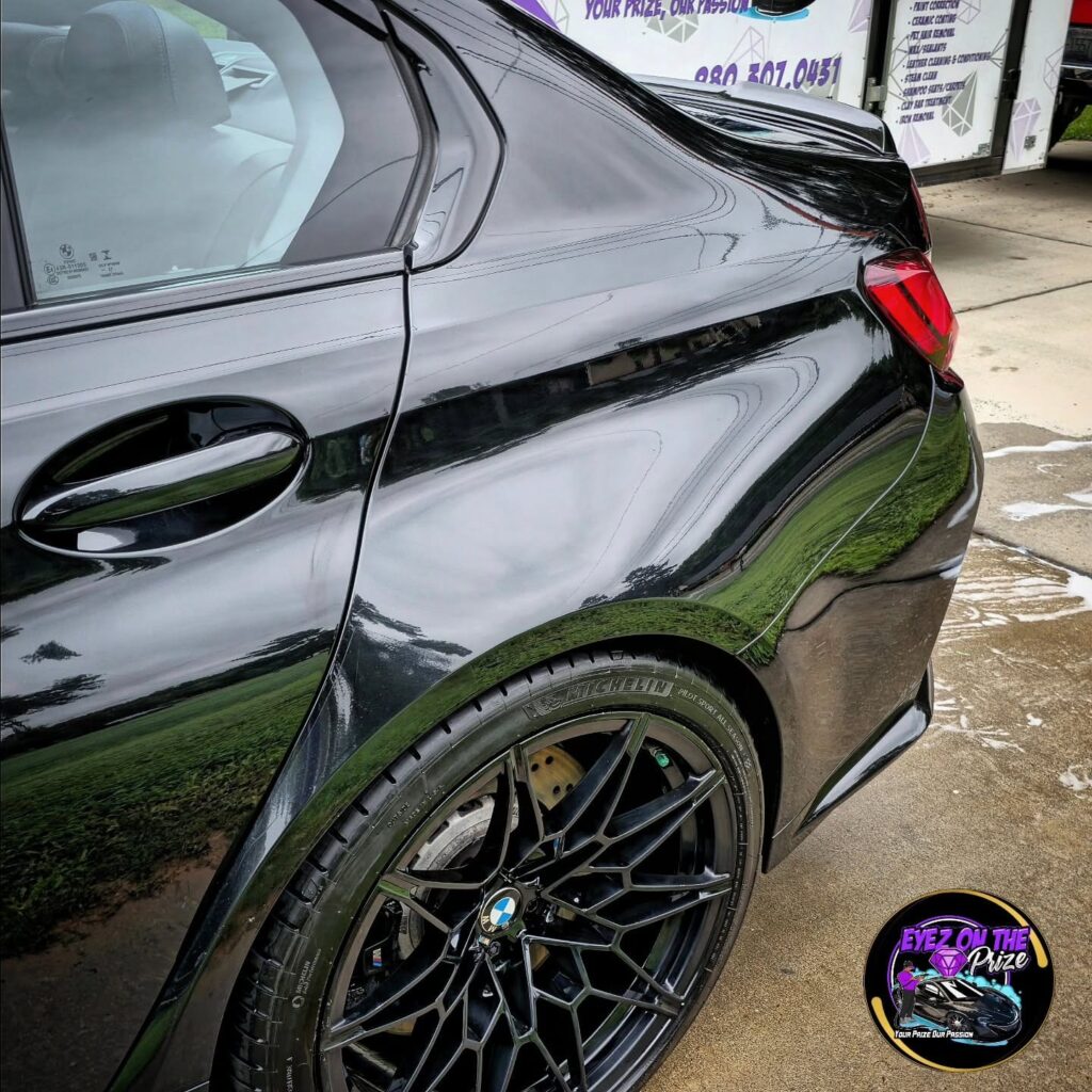 Rear quarter panel and black wheel of a glossy black BMW M3 parked beside a branded detailing trailer