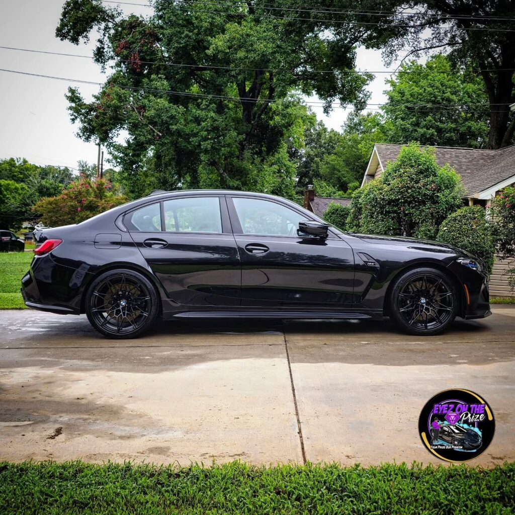 Side profile of a black BMW M3 with black wheels parked on a wet driveway after detailing