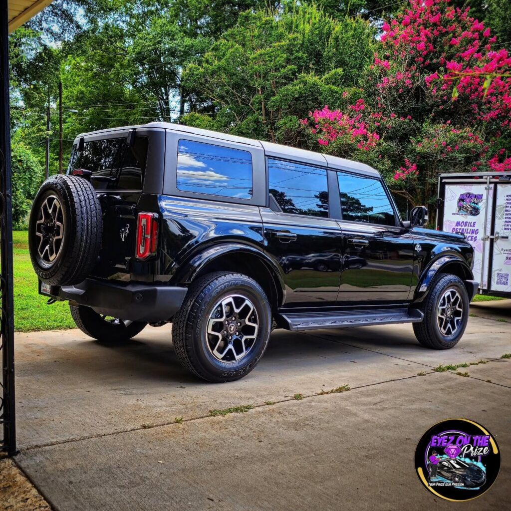 Black four-door Ford Bronco parked in a driveway after detailing, with a mobile detailing trailer in the background in