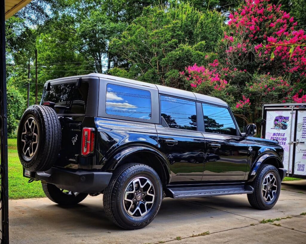 Black four-door Ford Bronco parked in a driveway after detailing, with a mobile detailing trailer in the background in