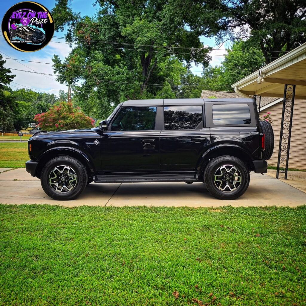 Side profile of a black four-door Ford Bronco with glossy paint parked on a residential concrete driveway