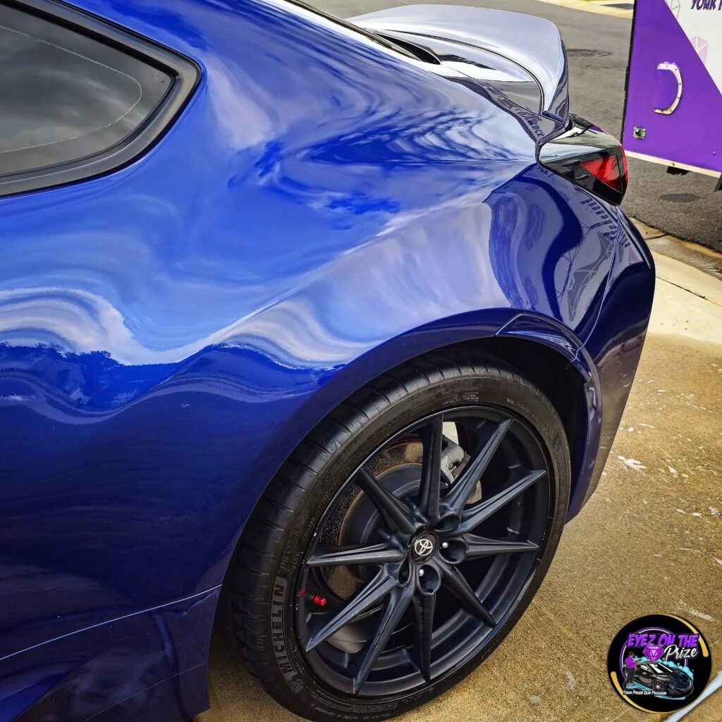 Rear quarter panel of a blue Toyota GR86 with glossy paint and black wheel, mobile detailing trailer in background