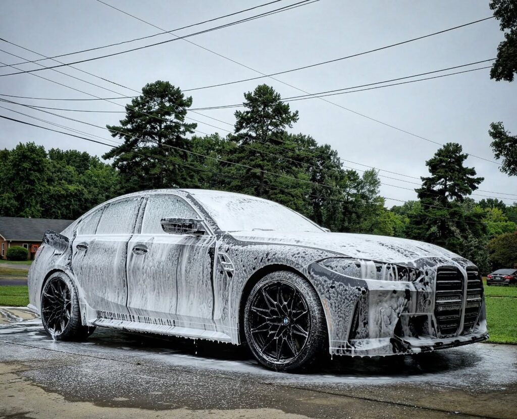 BMW M3 sedan covered in thick white foam during an exterior wash on a residential driveway in Charlotte, NC.