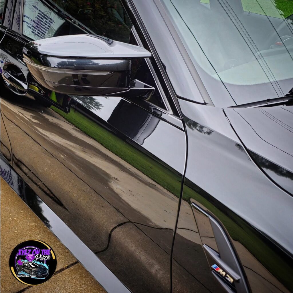 Close-up of a black BMW M3 side mirror, fender vent, and M3 badge with glossy reflective paint