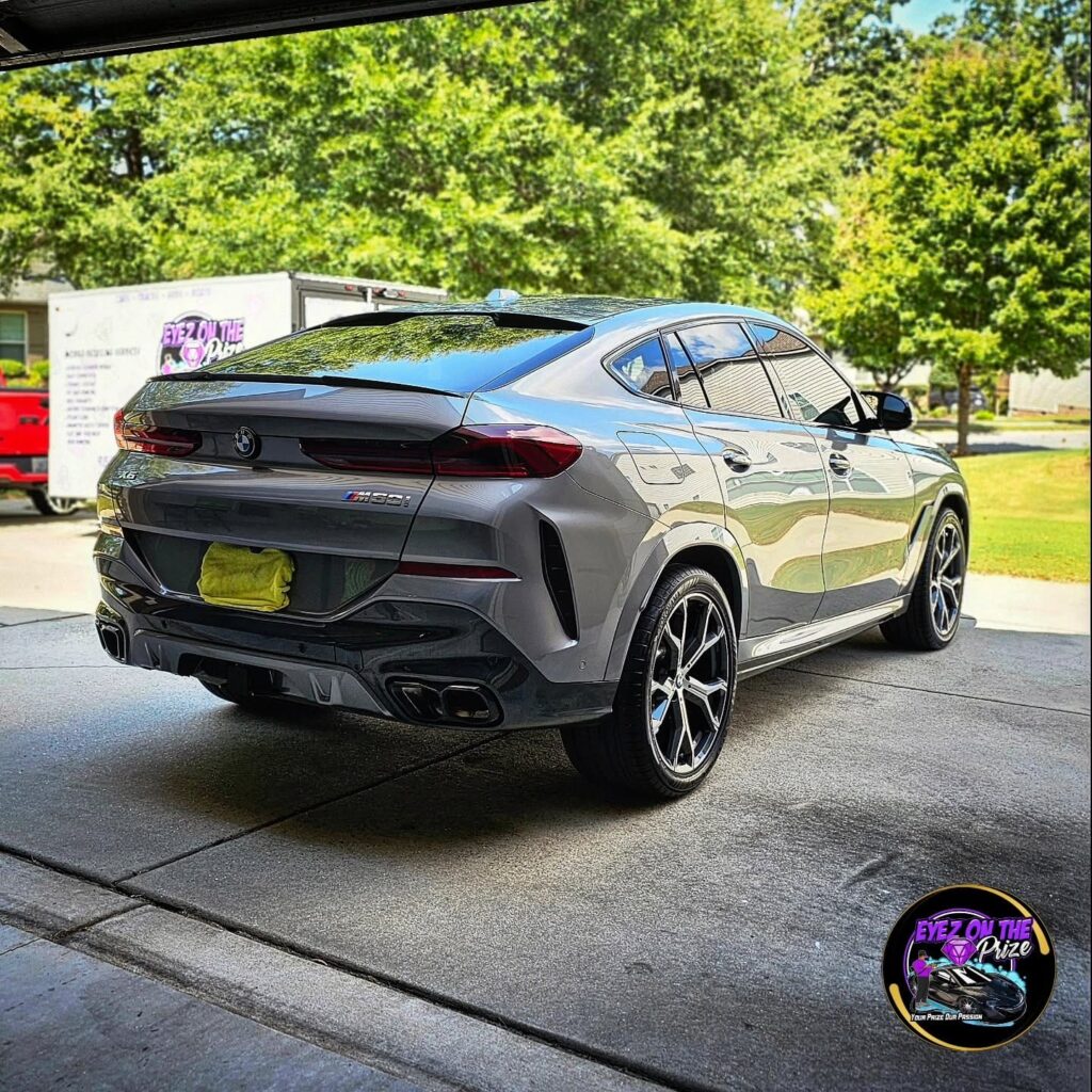 Rear quarter view of a gray BMW X6 M50i in a driveway with an Eyez On The Prize mobile detailing trailer behind it