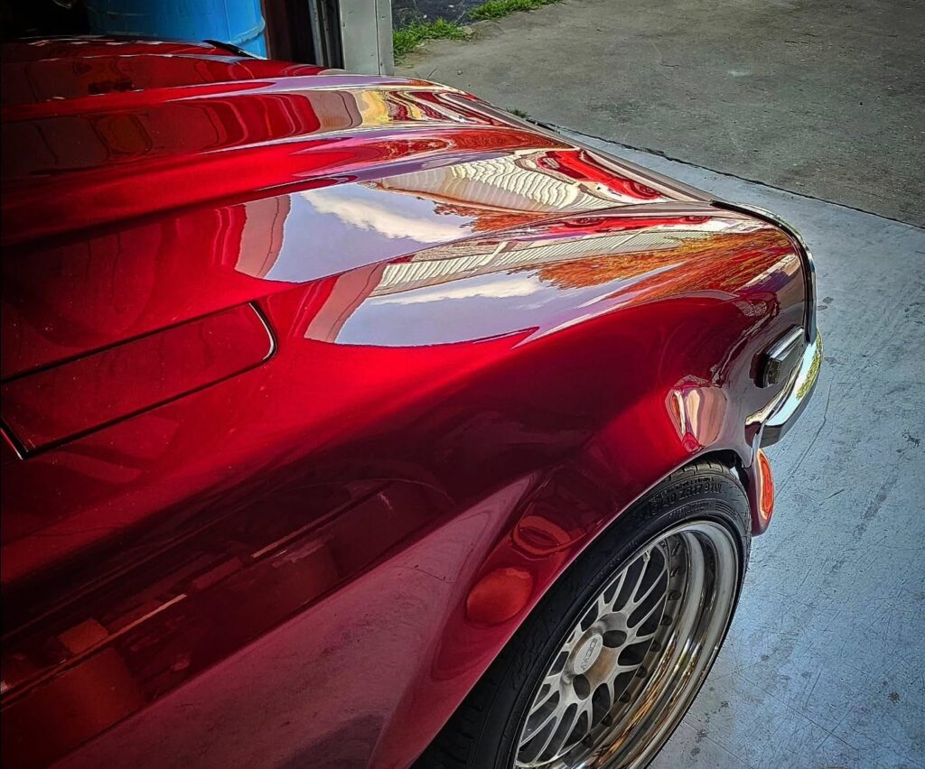 Close-up of a candy red classic car hood and front fender with a mirror-like polished finish and chrome multi-spoke wheel.