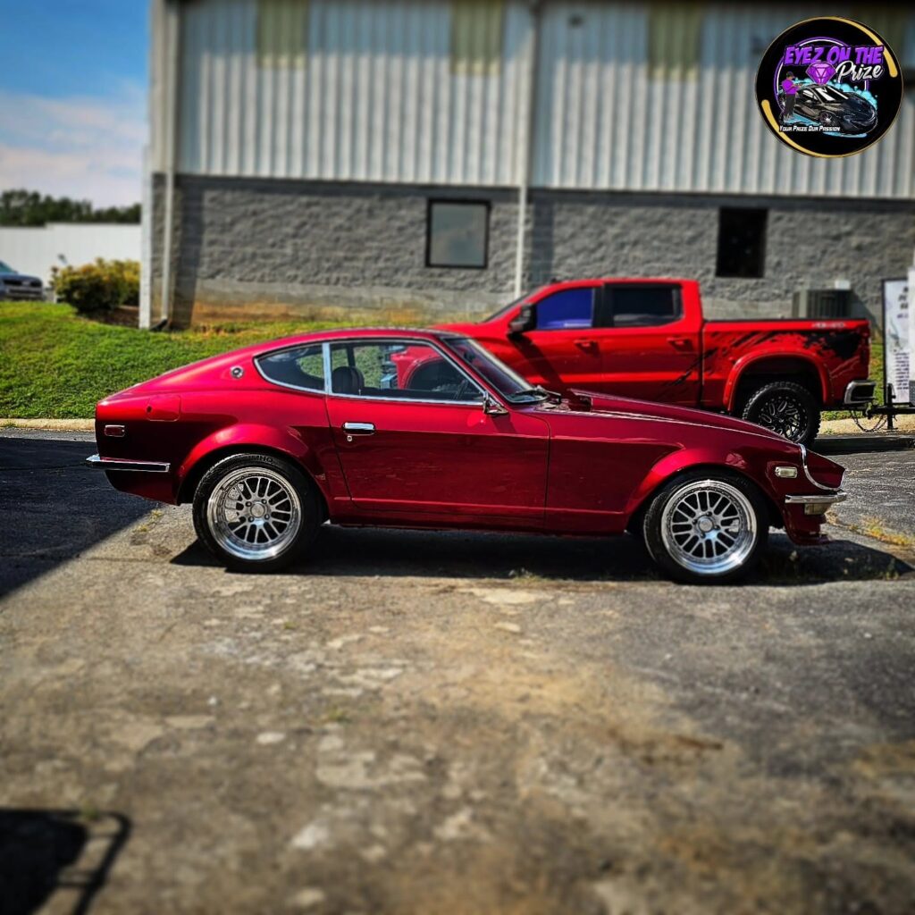 Side profile of a candy red Datsun 240Z with polished chrome wheels parked on pavement outside a building