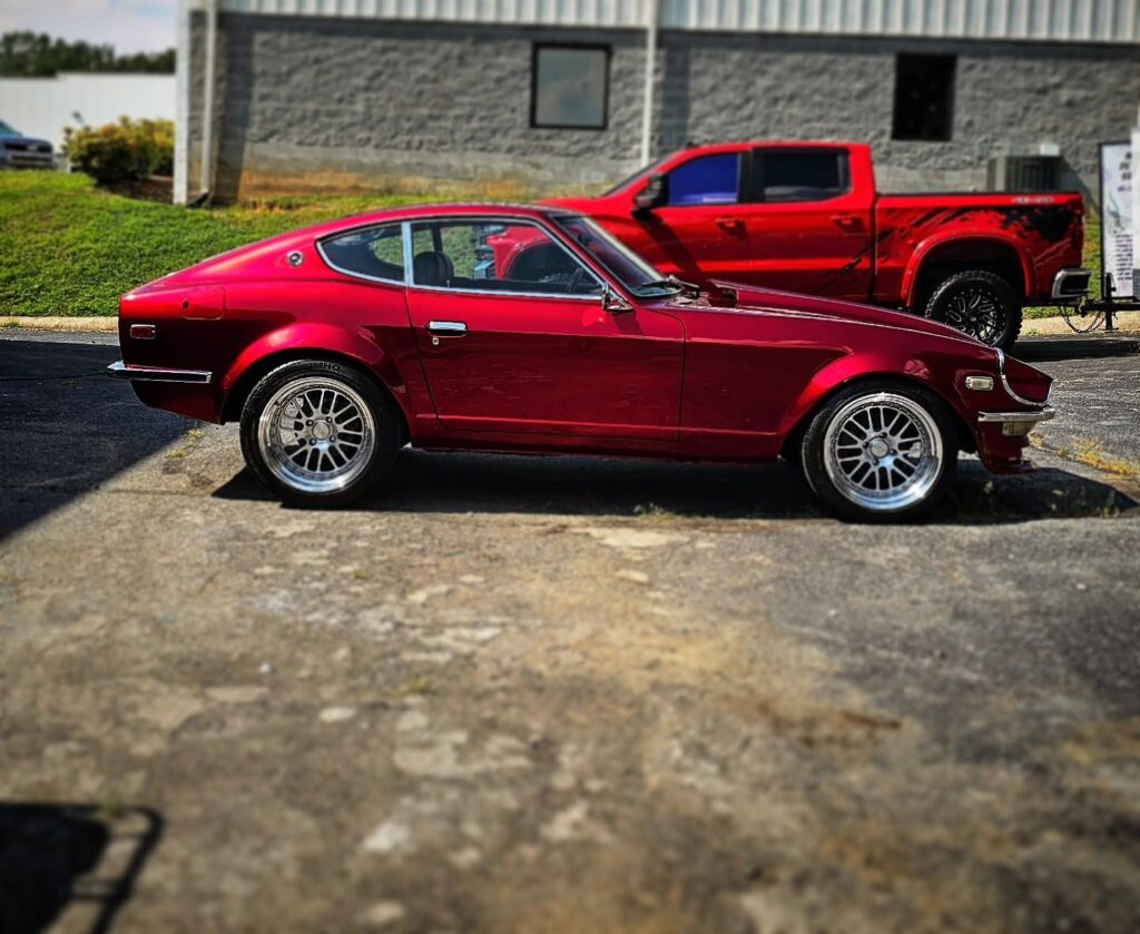 Side profile of a candy red Datsun 240Z with polished chrome wheels parked on pavement outside a building