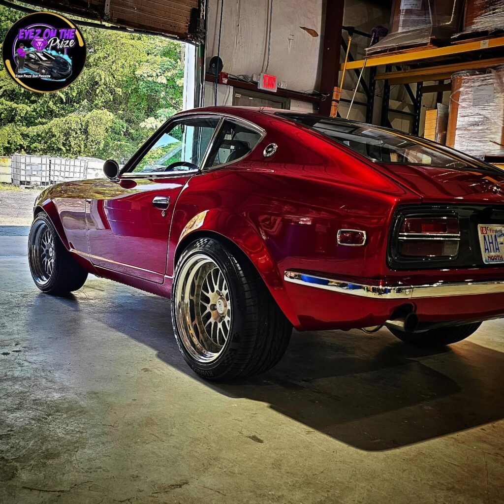 Rear quarter view of a candy red classic Datsun Z coupe with polished paint and aftermarket wheels inside a garage.