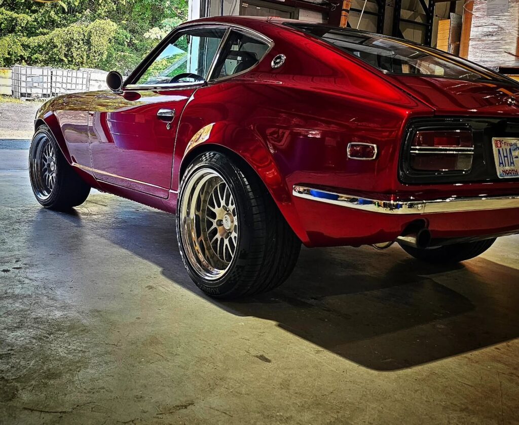 Rear quarter view of a candy red classic Datsun Z coupe with polished paint and aftermarket wheels inside a garage.