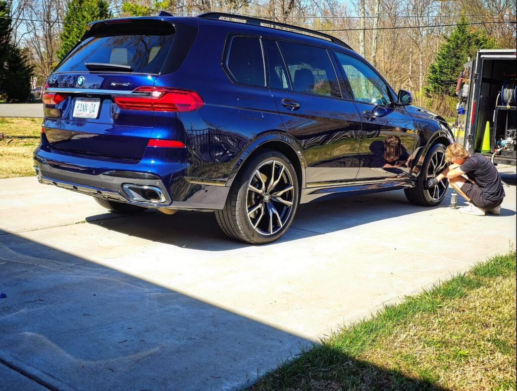 Dark blue BMW X7 in a driveway while a detailer works on the rear wheel next to a mobile detailing trailer.