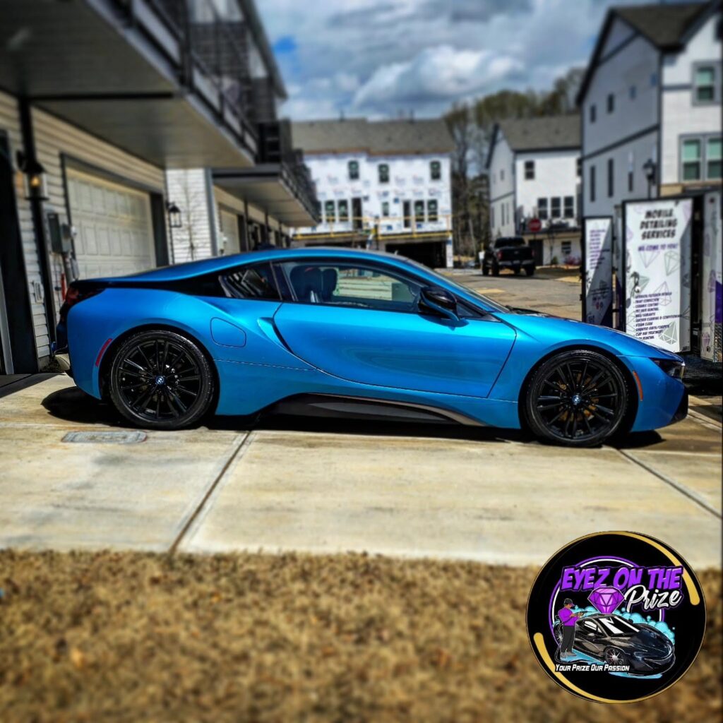 Blue BMW i8 parked on a driveway beside a garage, with a branded mobile detailing trailer in the background