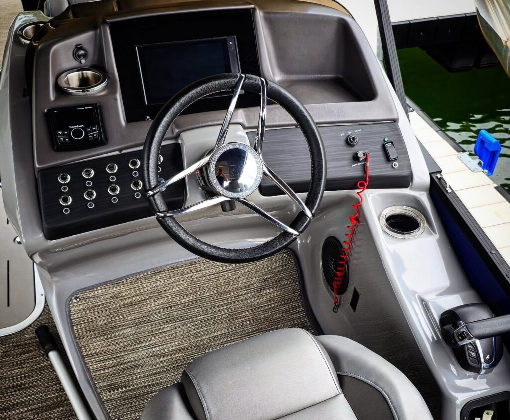 Cleaned pontoon boat helm with chrome steering wheel, gray captain's seat, and gauge console at a dock.