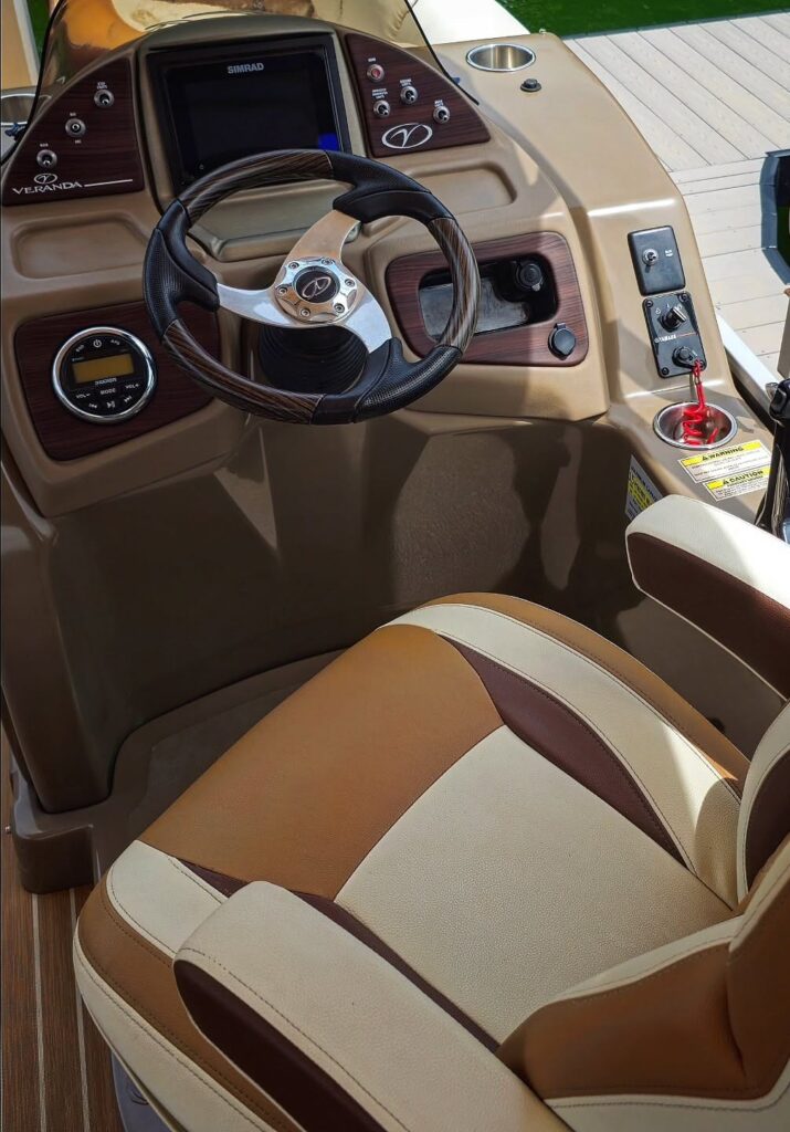 Clean helm of a Veranda pontoon boat with tan vinyl captain seat, wood-trim steering wheel, and Simrad display at a dock.