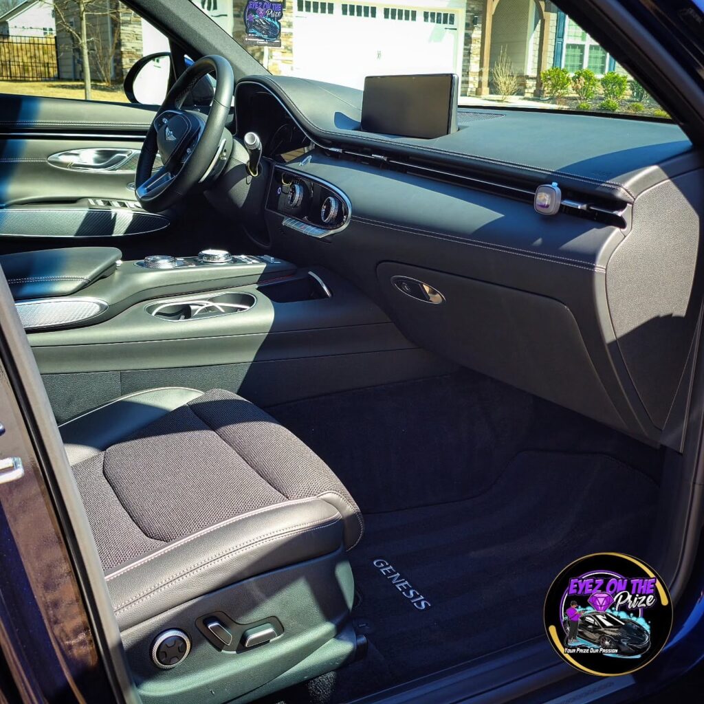 Clean Genesis GV70 interior seen through open passenger door, showing dashboard, front seat, and Genesis floor mat.