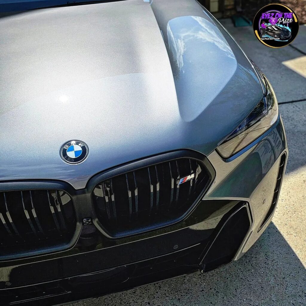 Close-up of a gray BMW X6 M hood, roundel badge, and black kidney grille with a glossy reflective finish.