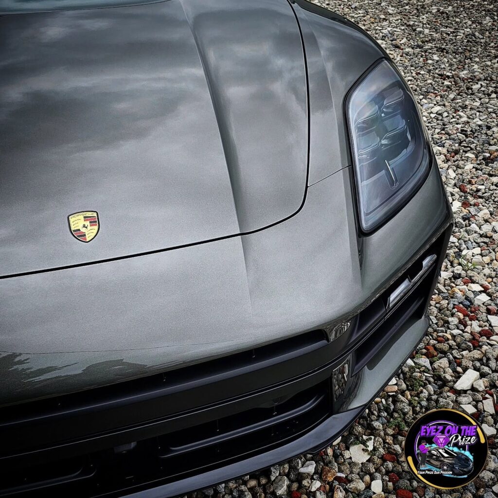 Close-up of a gray Porsche Macan hood, crest badge, and headlight showing a glossy reflective finish on gravel.
