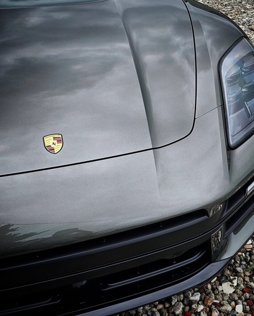 Close-up of a gray Porsche Macan hood, crest badge, and headlight showing a glossy reflective finish on gravel.