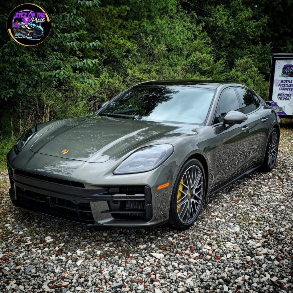 Gray Porsche Panamera with yellow brake calipers parked on gravel after detailing, mobile detailing trailer in background