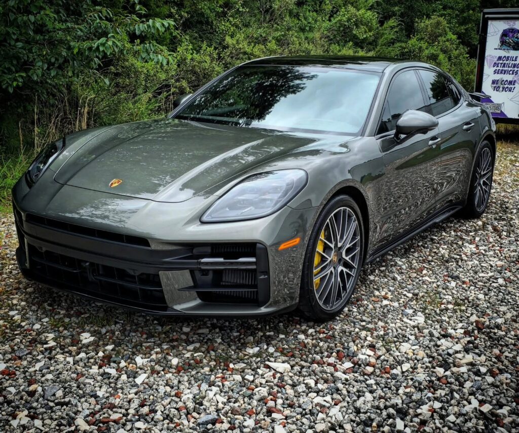 Gray Porsche Panamera with yellow brake calipers parked on gravel after detailing, mobile detailing trailer in background