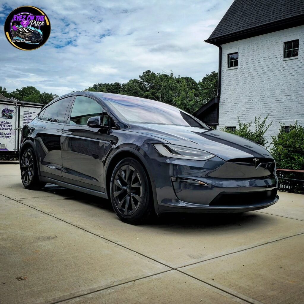 Gray Tesla Model X parked on a concrete driveway after detailing, with a mobile detailing trailer in the background