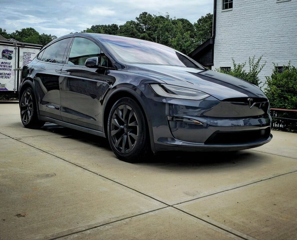 Gray Tesla Model X parked on a concrete driveway after detailing, with a mobile detailing trailer in the background