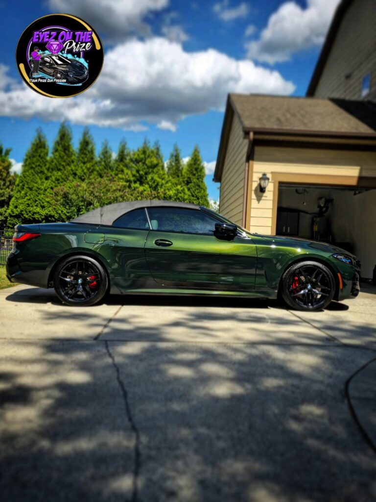 Side profile of a dark green BMW 4 Series convertible with black soft top parked on a driveway in front of a garage
