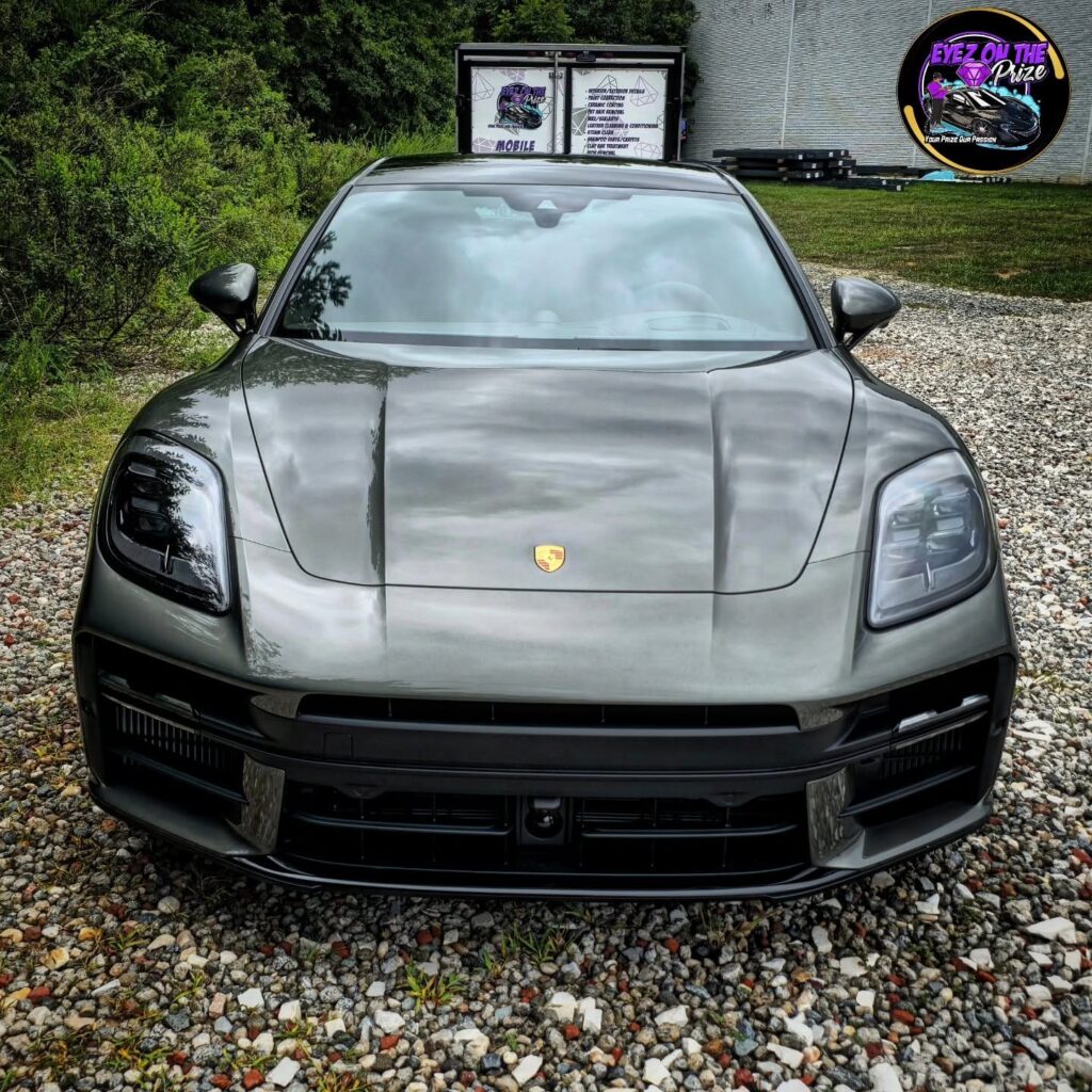 Front view of a gray Porsche Panamera with glossy hood parked on gravel, mobile detailing trailer signage in the background