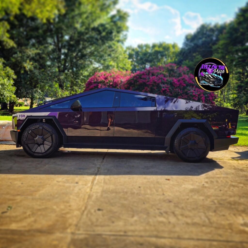 Side profile of a dark purple Tesla Cybertruck with glossy panels parked on a concrete driveway with trees behind