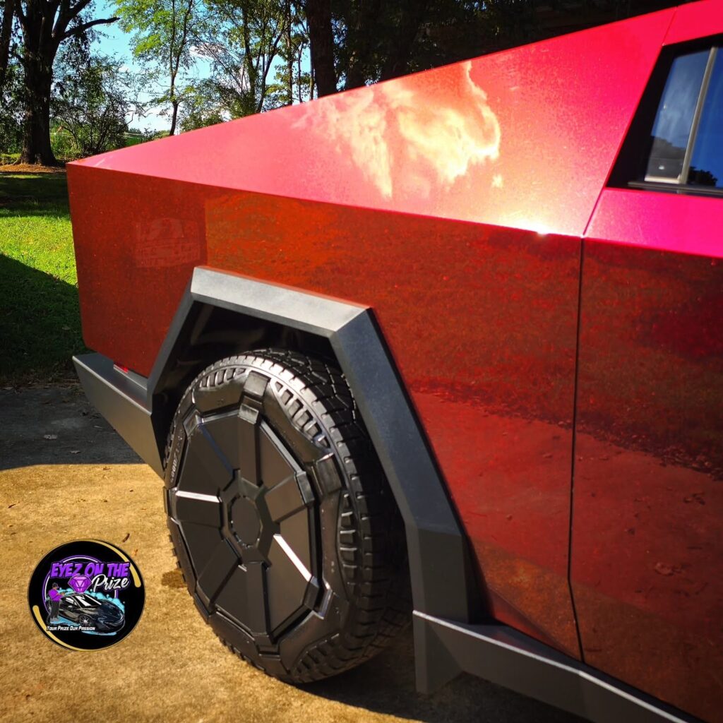 Close-up of a red Tesla Cybertruck rear quarter panel and black wheel with glossy paint reflecting trees and sky.