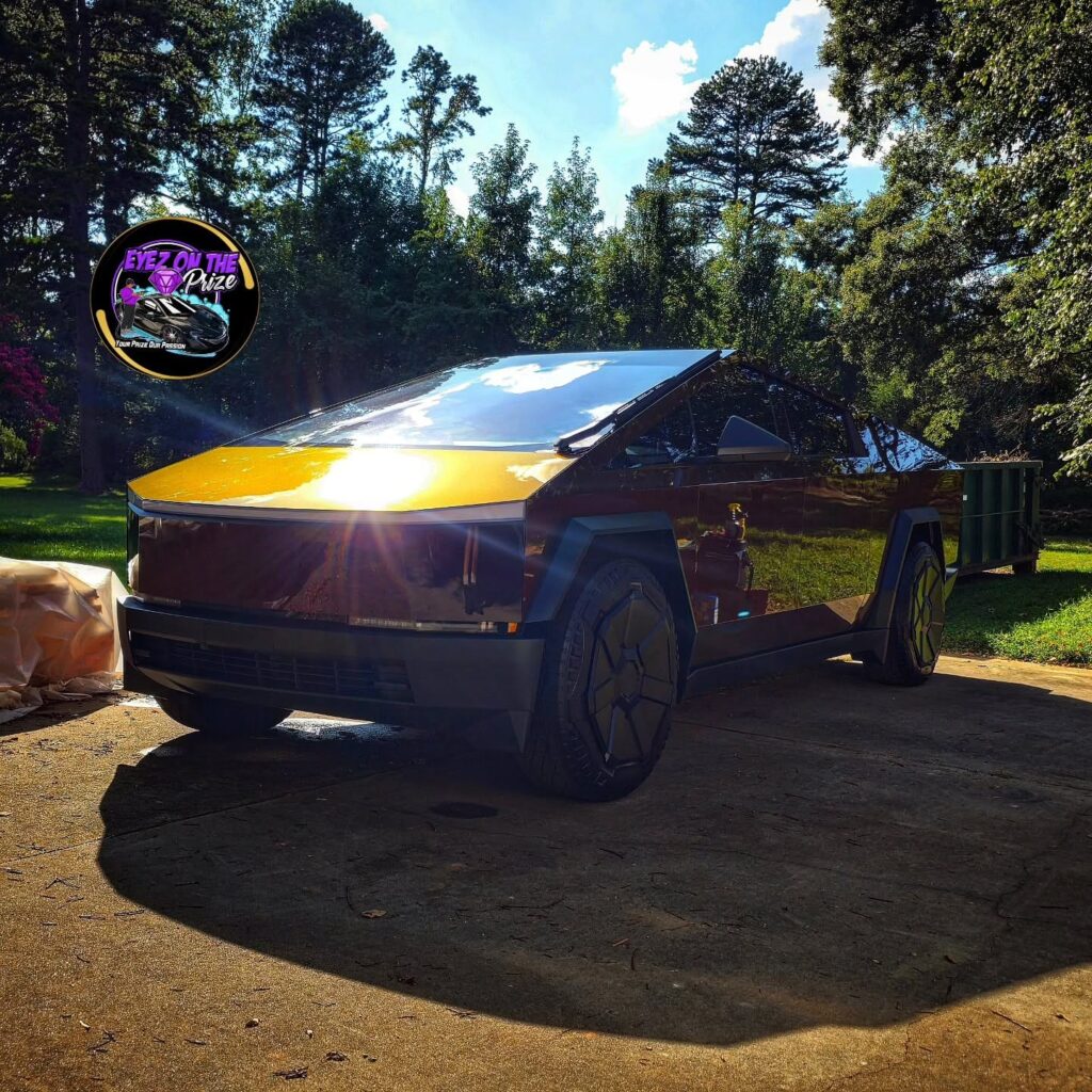 Tesla Cybertruck parked on a driveway with a glossy reflective finish, front quarter view in sunlight with trees behind