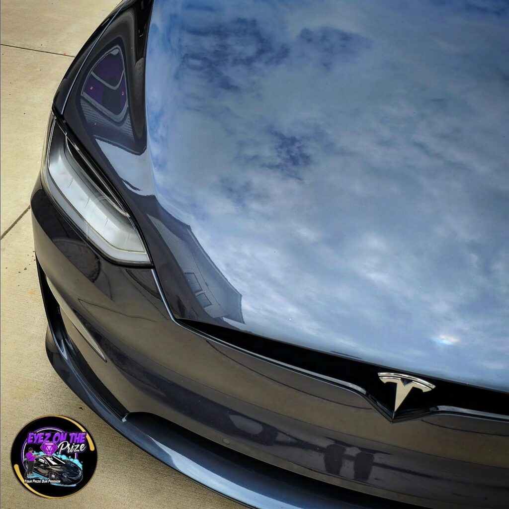 Close-up of a gray Tesla Model X hood and headlight with sky reflections in the polished paint after detailing.