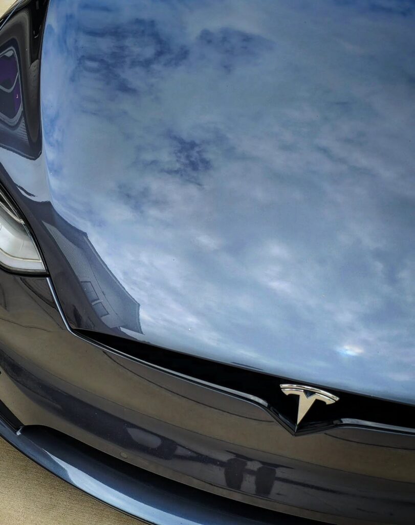 Close-up of a gray Tesla Model X hood and headlight with sky reflections in the polished paint after detailing.