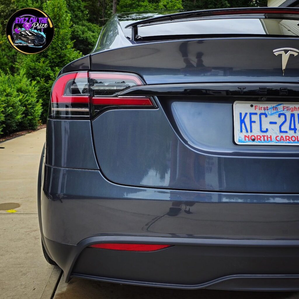 Close-up of a gray Tesla Model X rear end and taillight with a glossy finish, parked on a driveway near greenery.