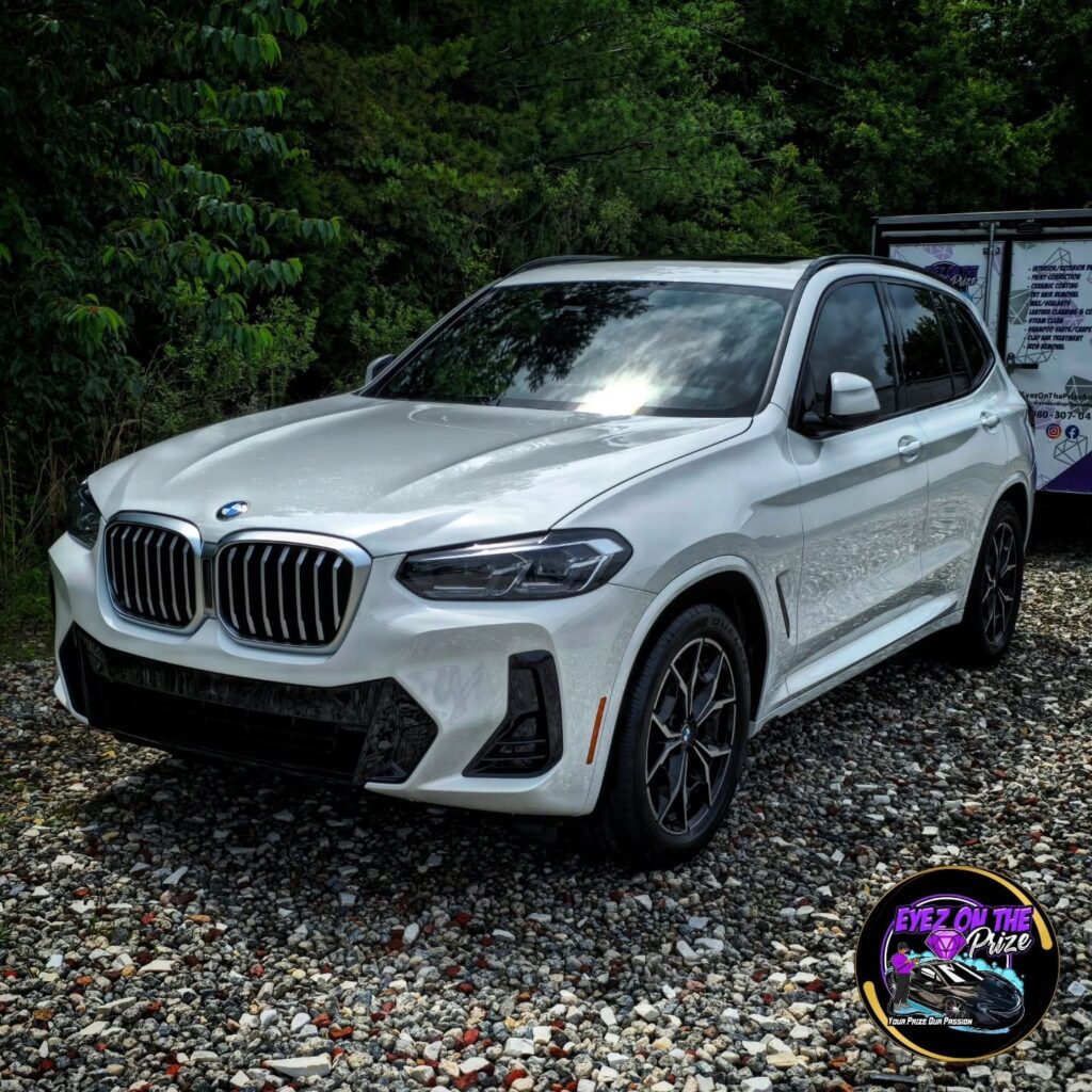 White BMW X3 front three-quarter view on a gravel lot with an Eyez On The Prize detailing trailer in the background