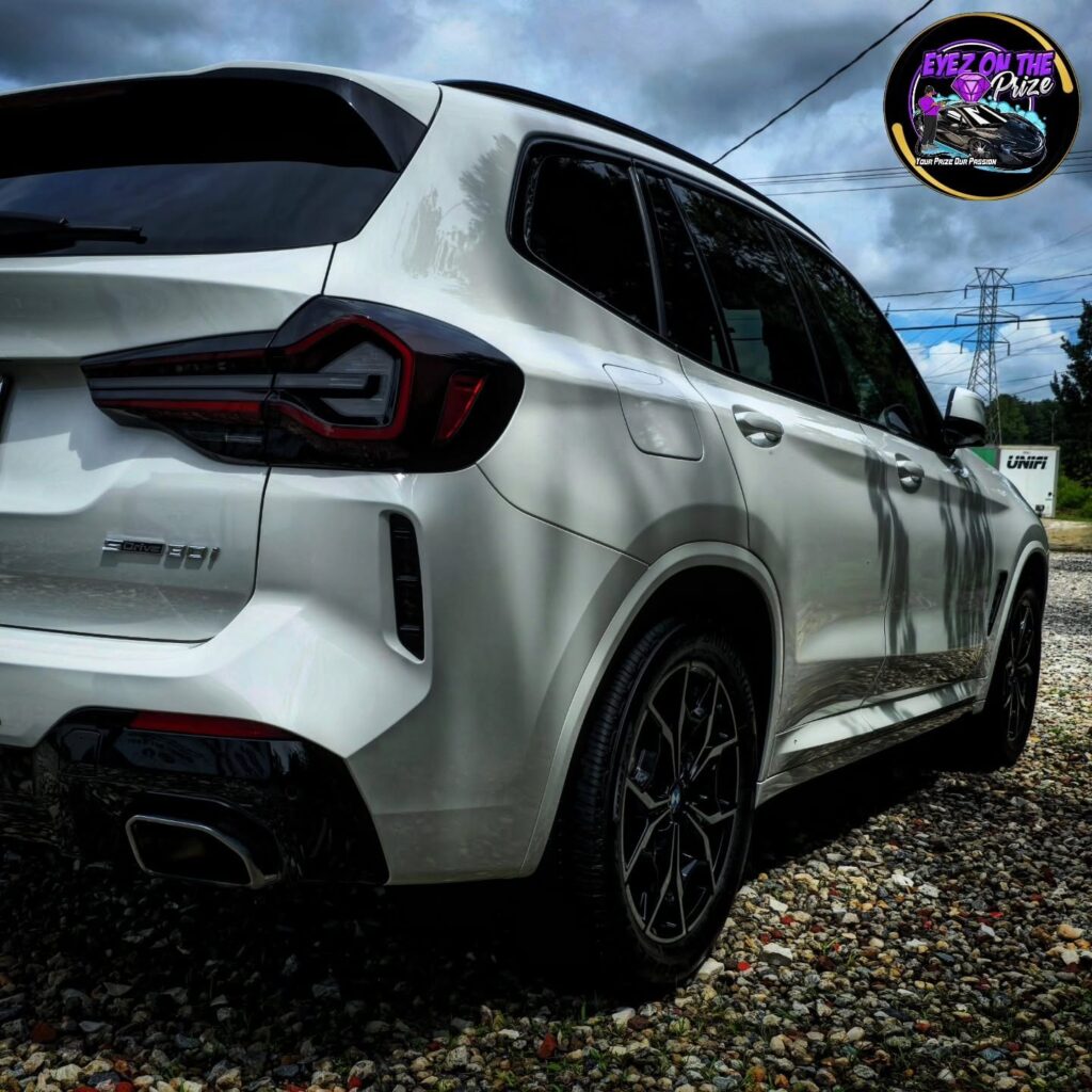 Rear quarter view of a white BMW X3 sDrive30i with clean paint and gloss black wheels, parked on gravel under cloudy skies.
