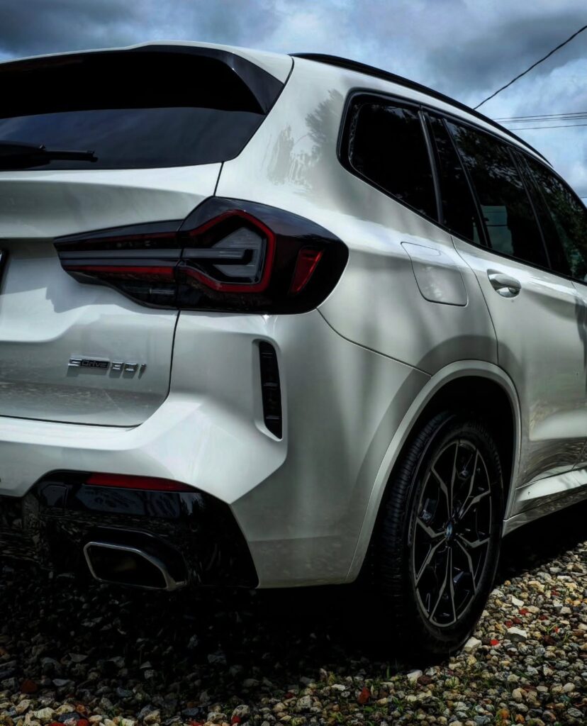 Rear quarter view of a white BMW X3 sDrive30i with clean paint and gloss black wheels, parked on gravel under cloudy skies.