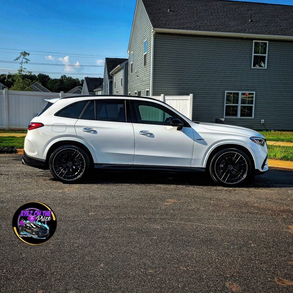 White Mercedes AMG GLC SUV with black multi-spoke wheels parked on a residential street beside a white fence and gray house.