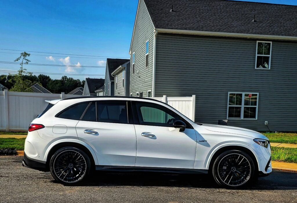 White Mercedes AMG GLC SUV with black multi-spoke wheels parked on a residential street beside a white fence and gray house.