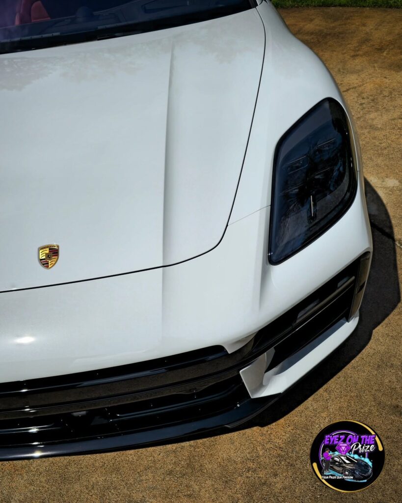 Close-up of a white Porsche hood and headlight with glossy paint and the Porsche crest, photographed outdoors.